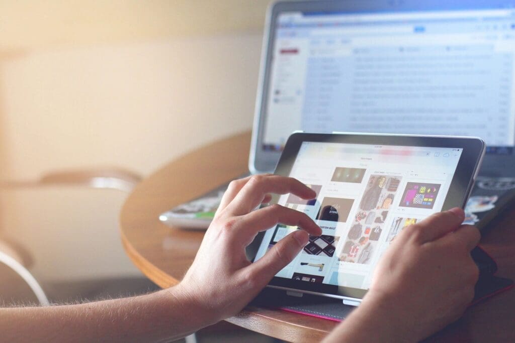 Hands browsing images on a tablet with a laptop in the background.