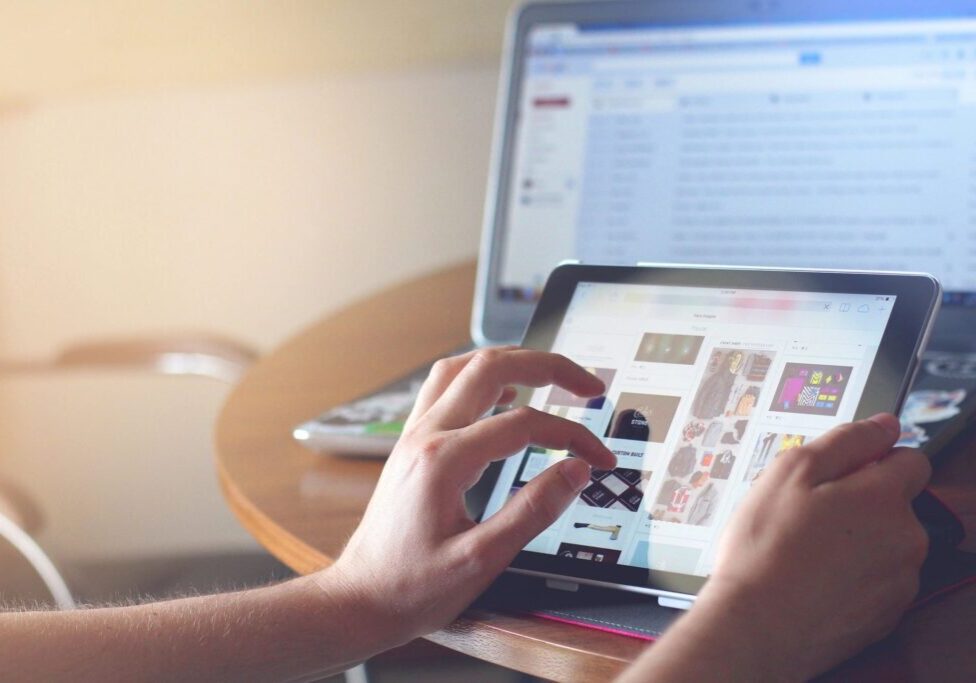 Hands browsing images on a tablet with a laptop in the background.